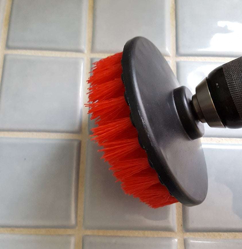 Red flat 5 inch cleaning drill brush with stiff nylon bristles attached to a drill scrubbing tile grout.